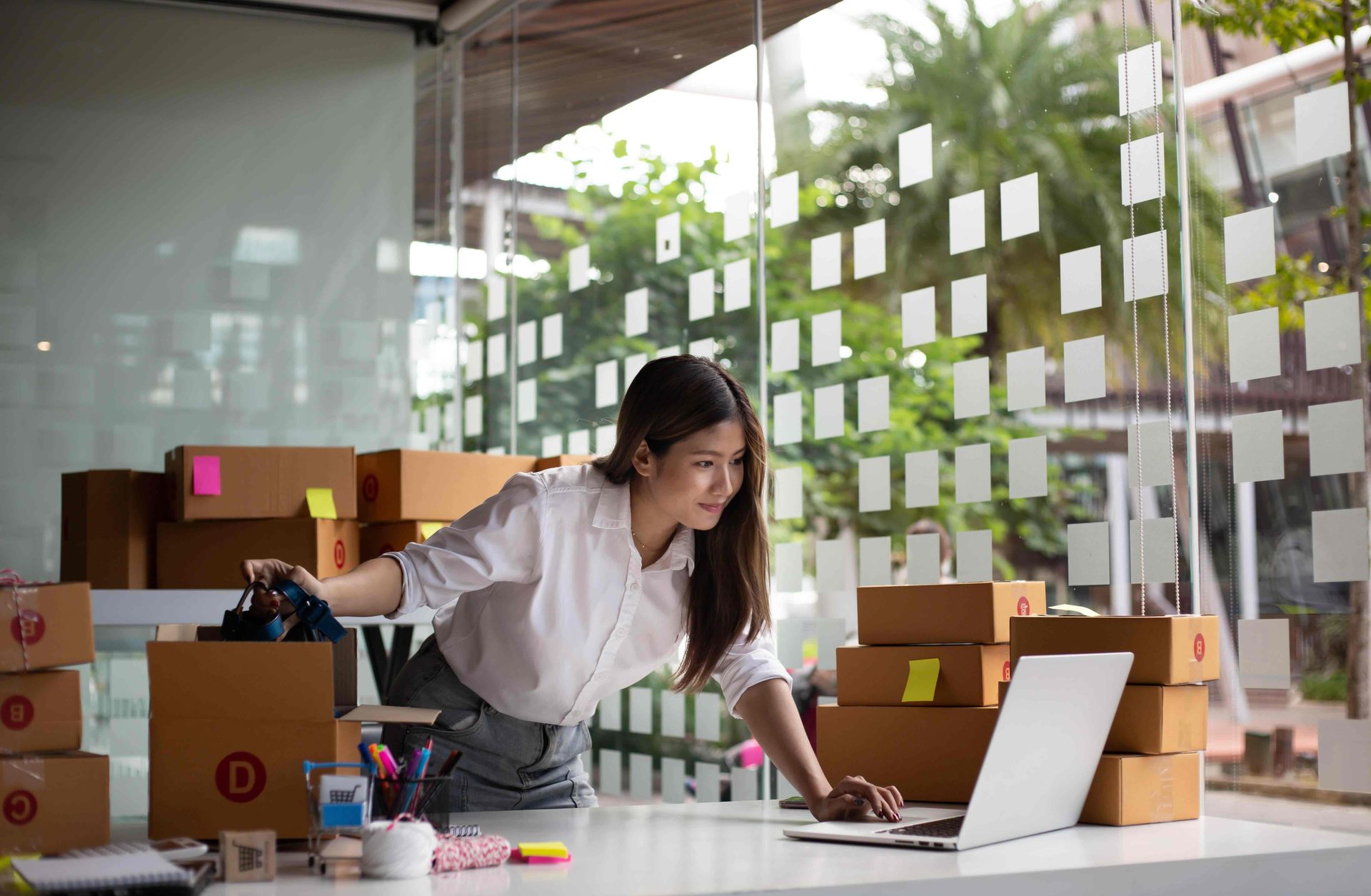 Woman leaning over desk with laptop, surrounded by stacked shipping boxes in office setting.