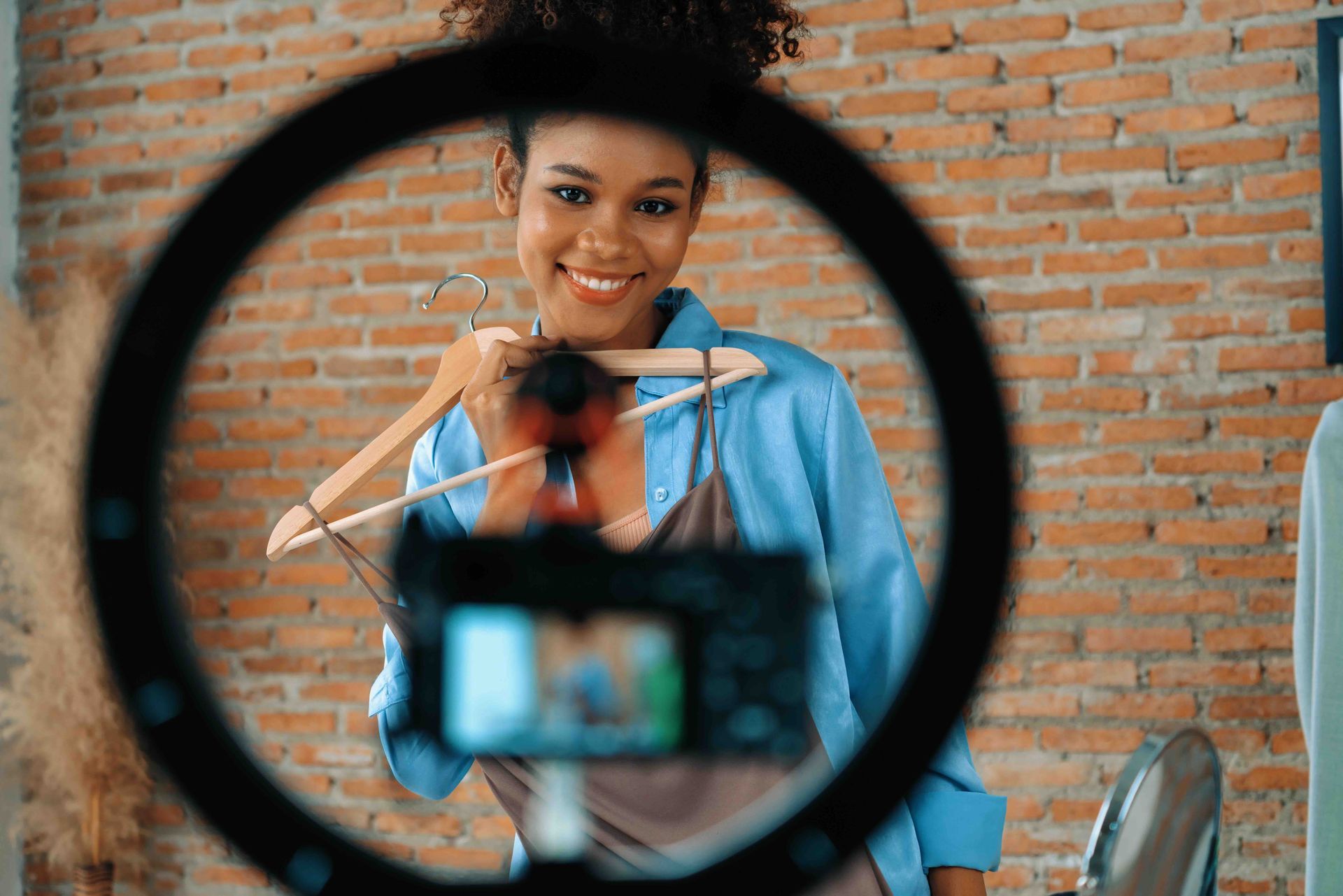 Woman in a blue shirt holding a garment on a hanger, smiling at a camera set up through a ring light, with a brick wall background.