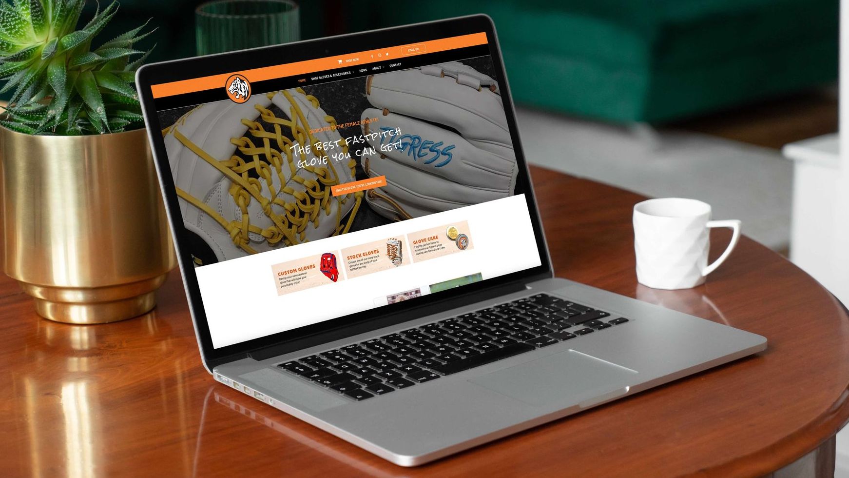 A laptop computer with the Tigress Gloves website on the sceren is sitting on a wooden table next to a cup of coffee.