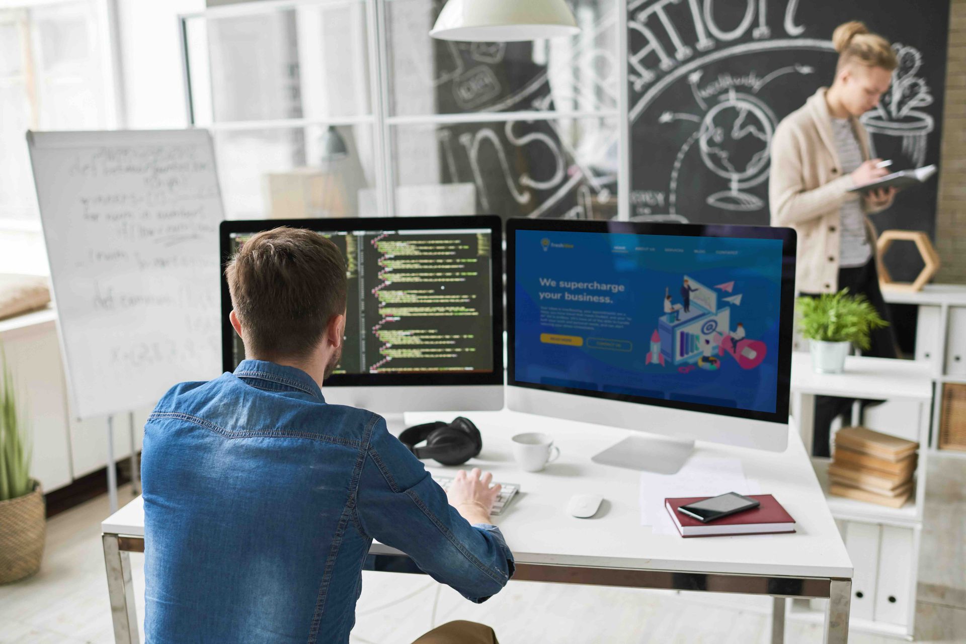 Man working on computer code with two monitors at desk; colleague in background.