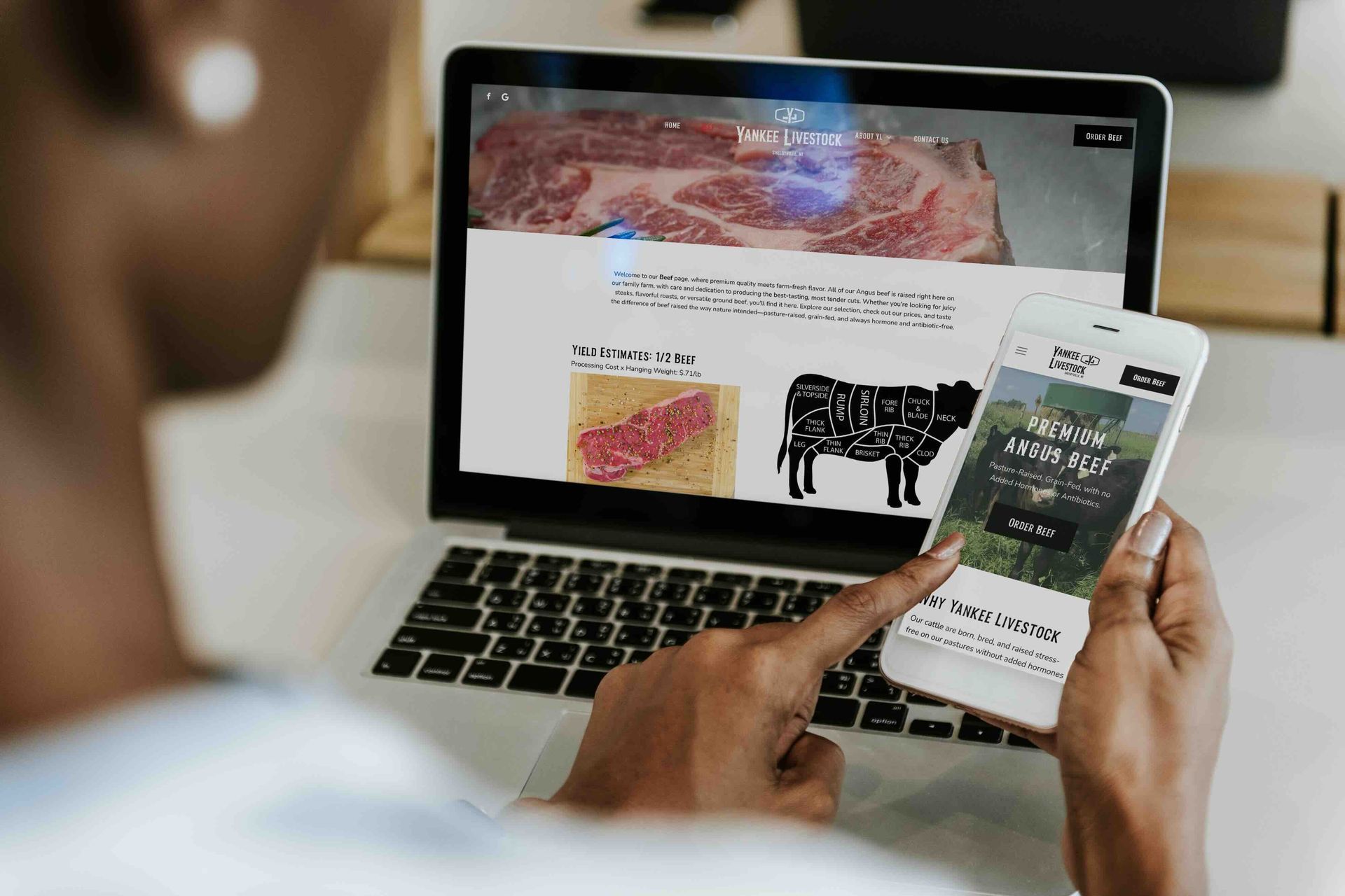 A person is holding a cell phone in front of a laptop which both show the Yankee Livestock website.