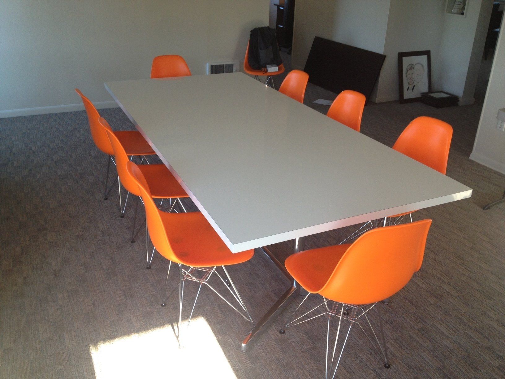 A long, light gray table with eight orange chairs in a bright room.