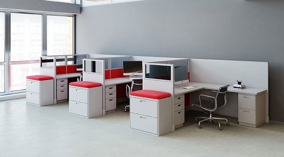 Office cubicles, white desks with red accents and stools, grey wall, large windows.