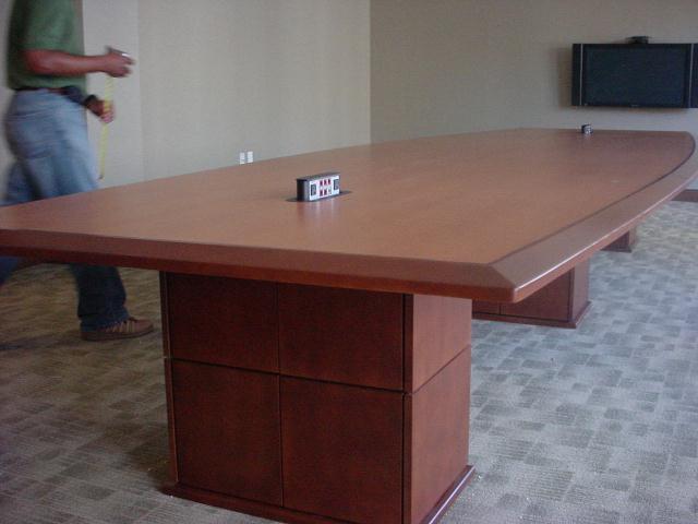 Man in blue jeans walking past a large wood conference table; level on table; TV on wall.