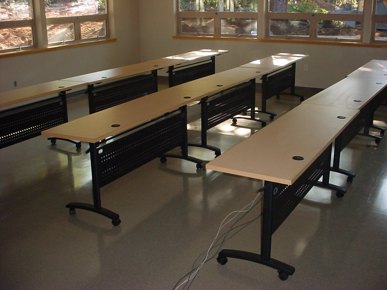 Long, rectangular desks in a classroom, with black frames and beige tops. Sunlight streams through windows.