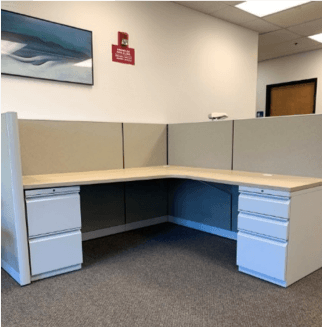 L-shaped office desk with beige cubicle walls, white drawers, and a gray carpet floor.