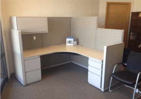 L-shaped office cubicle with beige desk, tan walls, and storage drawers. A black chair sits to the side.