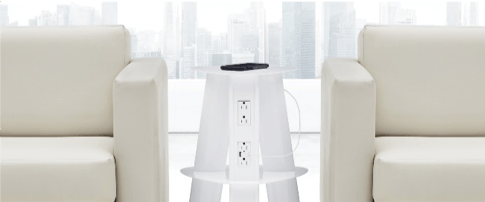 Two cream-colored armchairs flanking a transparent, white side table with outlets and a remote control on top, in front of a window.