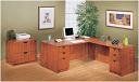 Wooden office with L-shaped desk, filing cabinets, computer, printer, and art; a green plant is in the background.