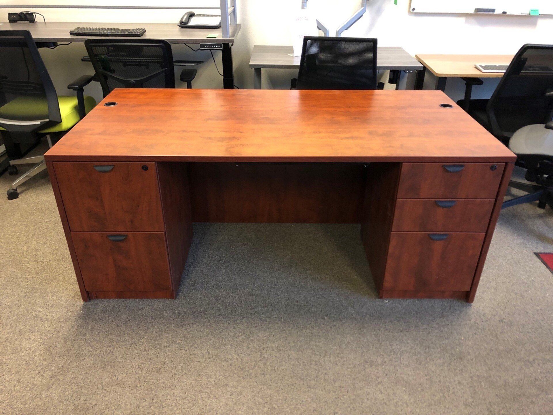 Wooden office desk with drawers, brown finish. Office setting, neutral carpet.