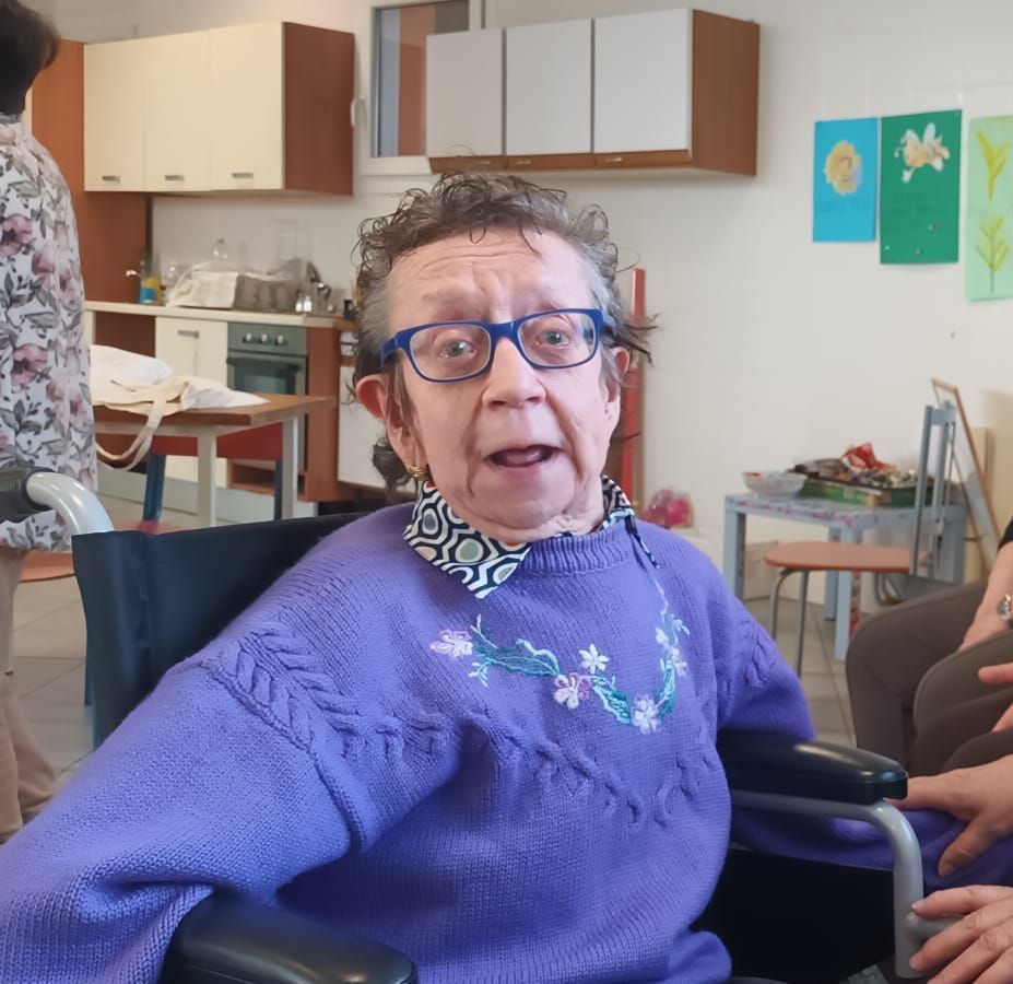 Una donna anziana con gli occhiali e un maglione viola è seduta su una sedia a rotelle