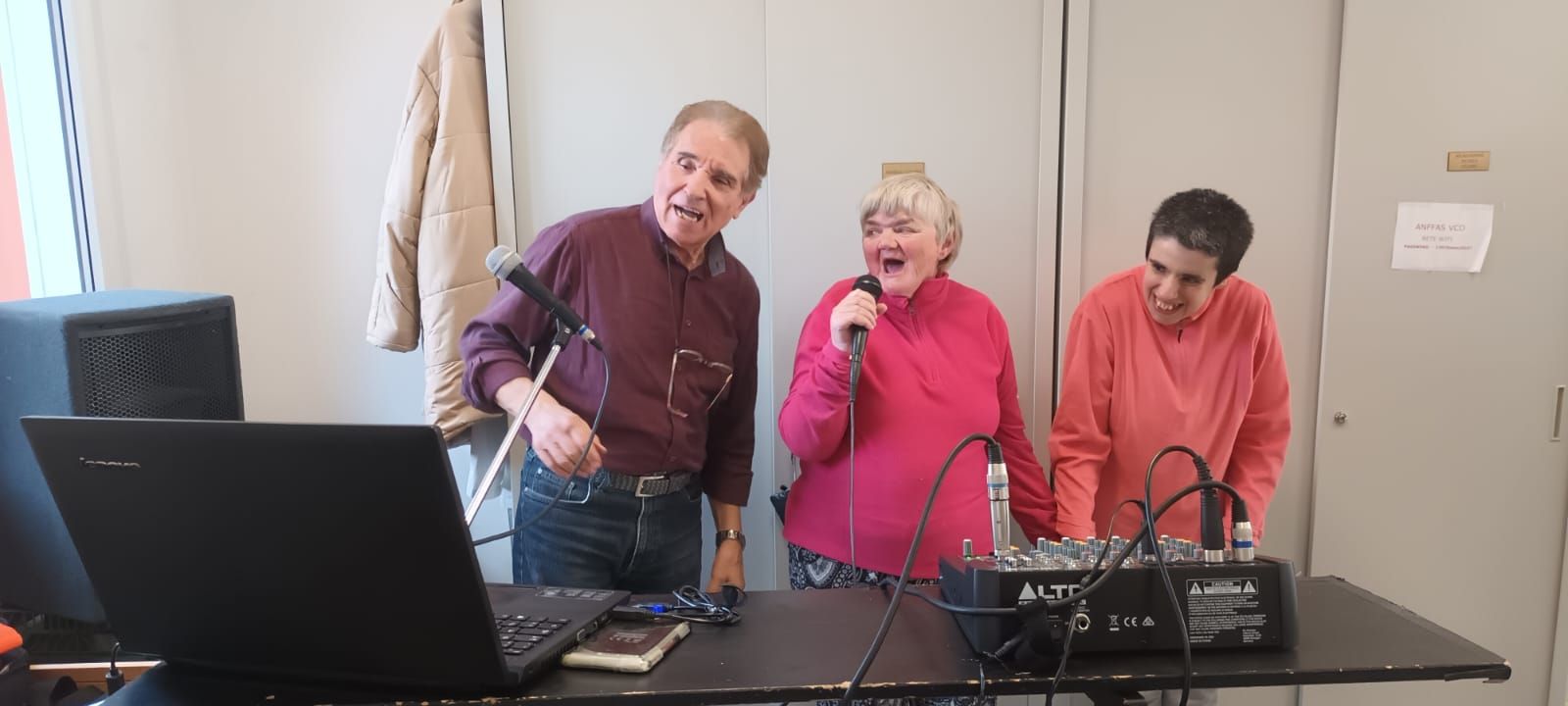 Un gruppo di persone è in piedi attorno a un tavolo e canta ai microfoni.