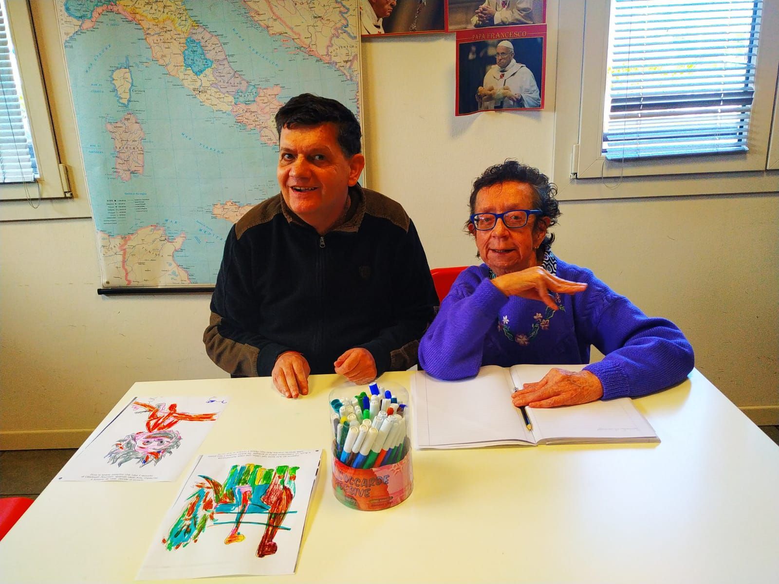 Due persone sono sedute a un tavolo con una mappa appesa al muro dietro di loro