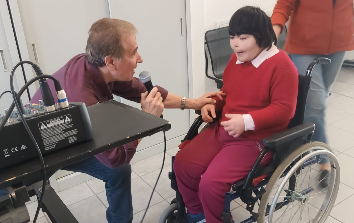 Un uomo parla al microfono mentre una ragazza su una sedia a rotelle guarda.