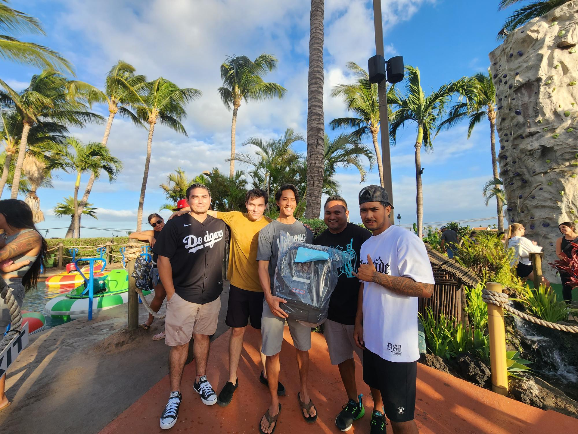 Five young men pose with a large object at a miniature golf course, surrounded by palm trees under a sunny sky.