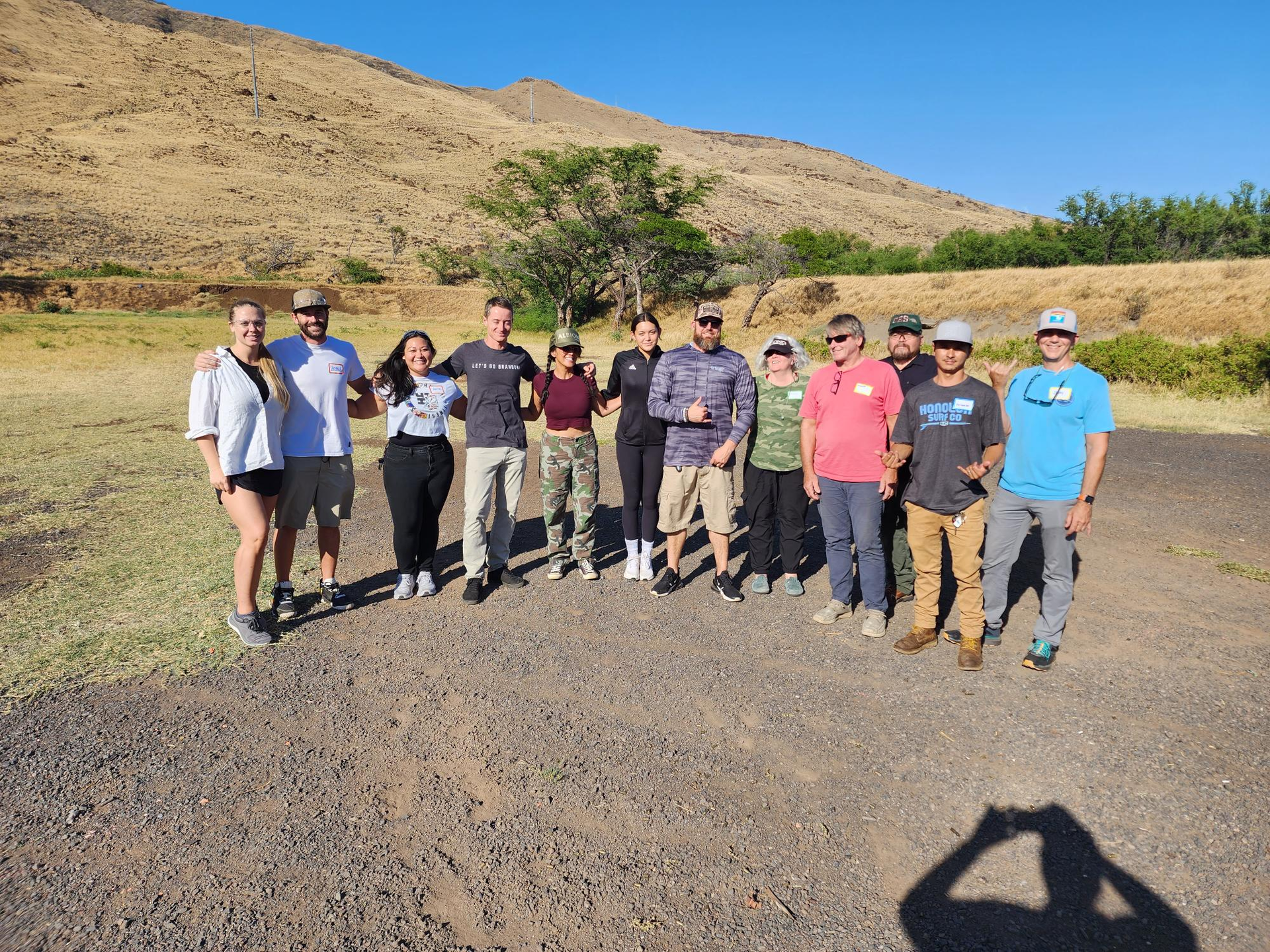 Group of people standing outdoors, posing for a photo. They are in front of a dry, hilly landscape on a sunny day.