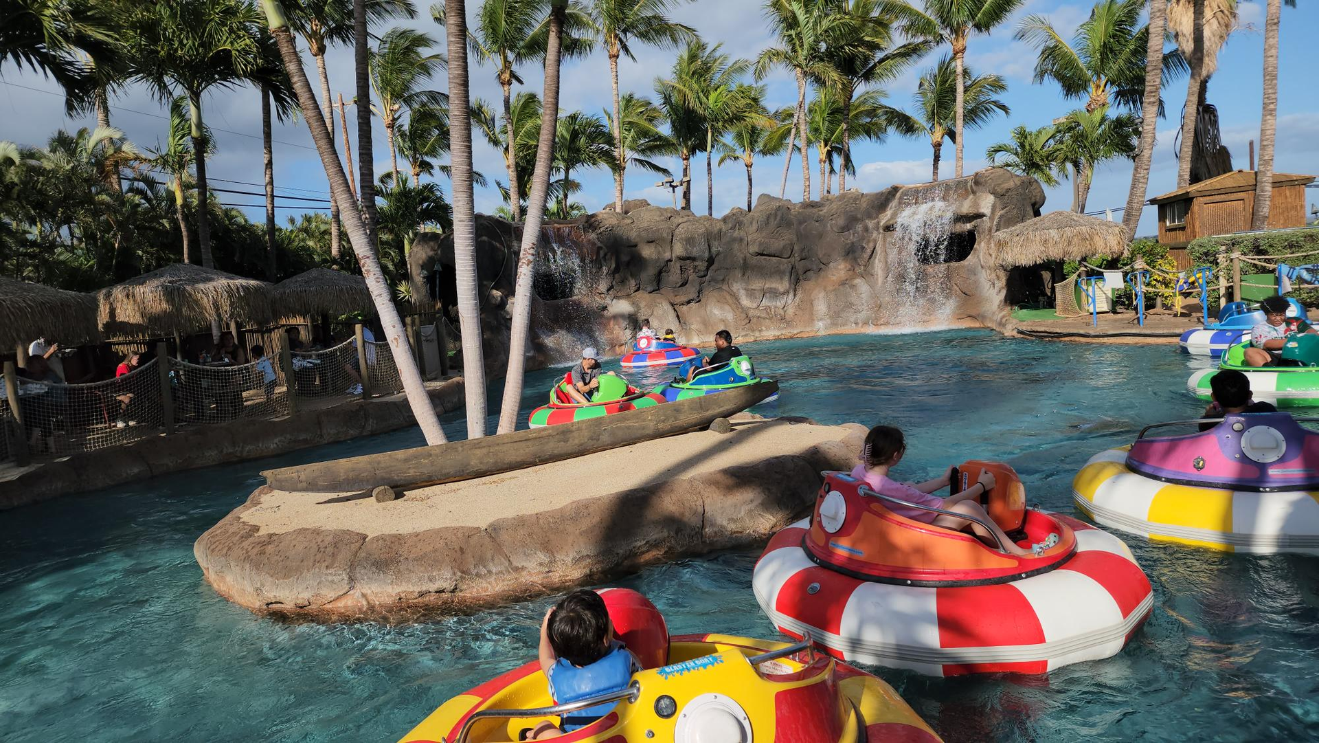 People in colorful bumper boats on a water ride with a waterfall feature and palm trees in the background.