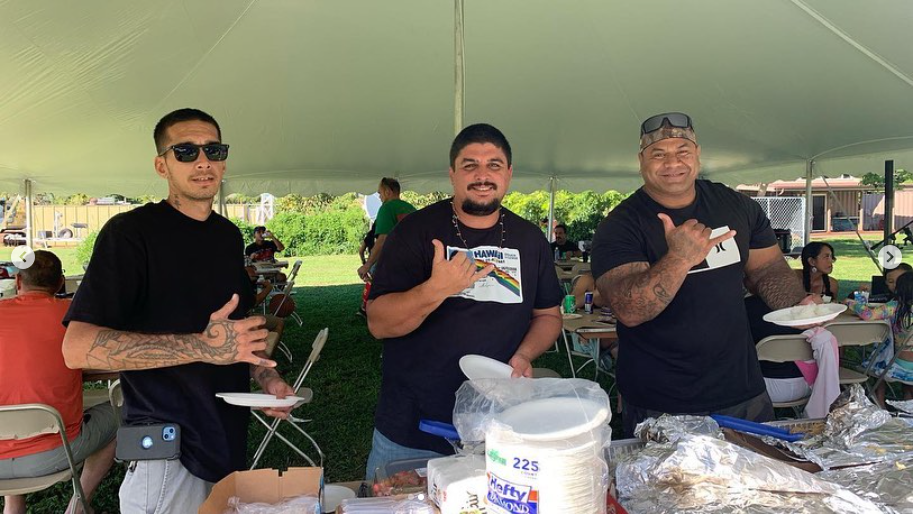 Three men at an outdoor event, under a tent, gesturing with hands. They are holding plates and appear to be serving food.