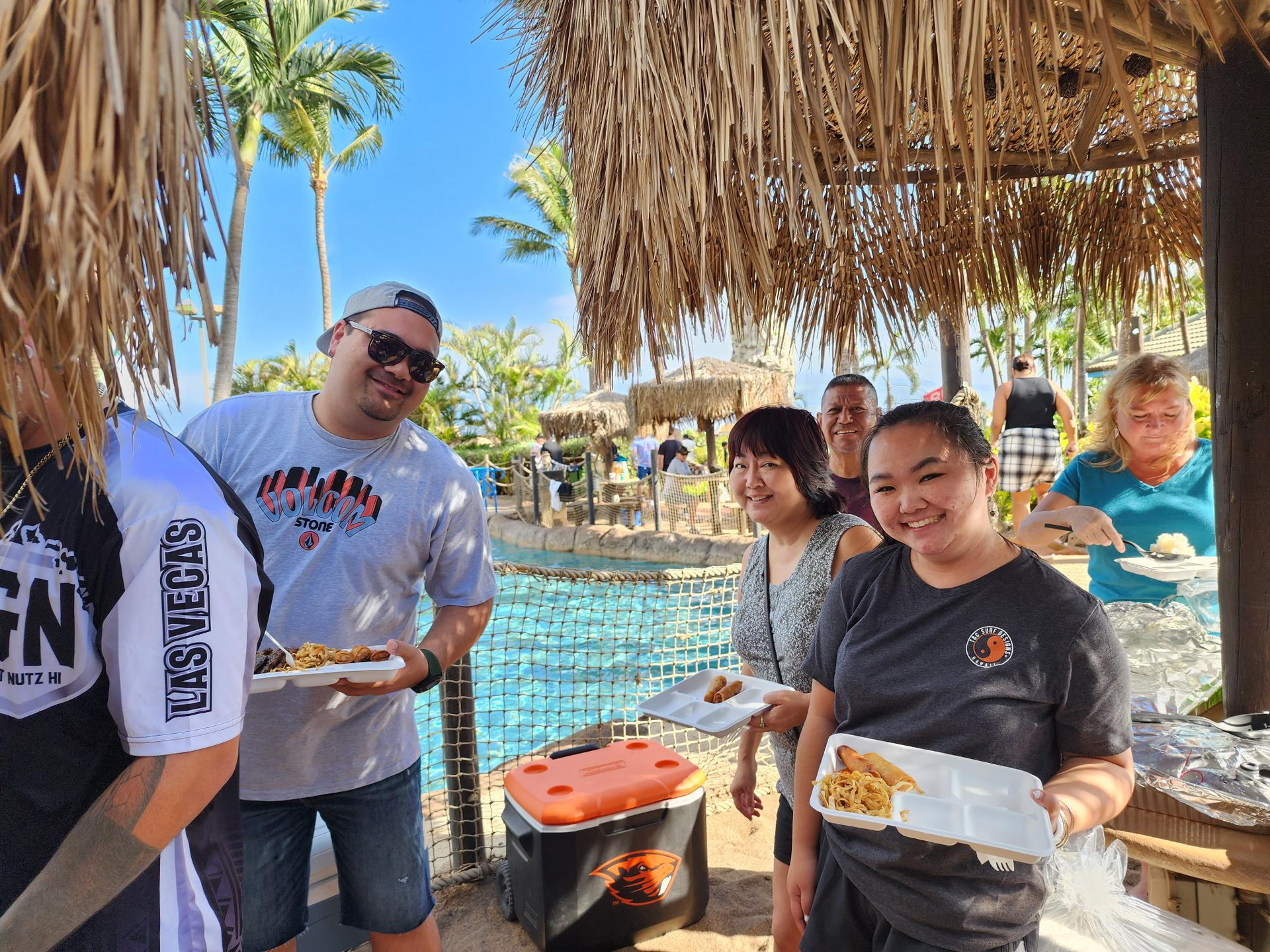A group of people at an outdoor event near a pool, holding plates of food. Sunny day, palm trees in the background.