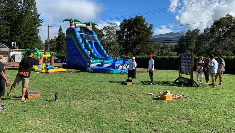 A group of people enjoying outdoor games on a grassy lawn. Inflatable water slide, cornhole, and other games set up