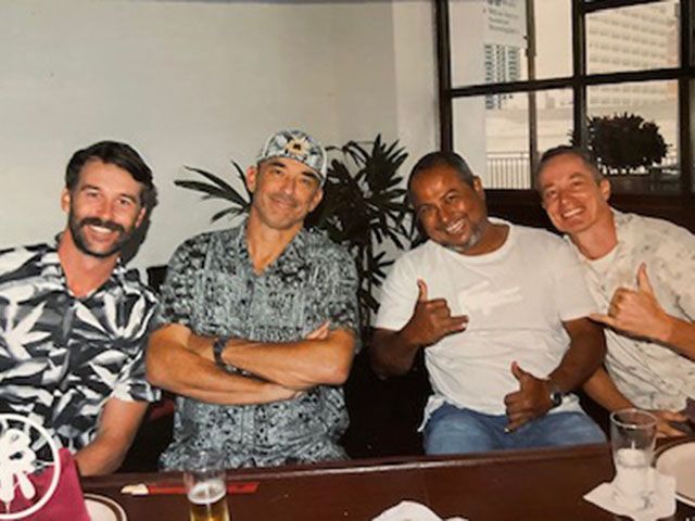 Four men smile for a photo at a restaurant. They wear casual shirts, one gives a shaka sign.