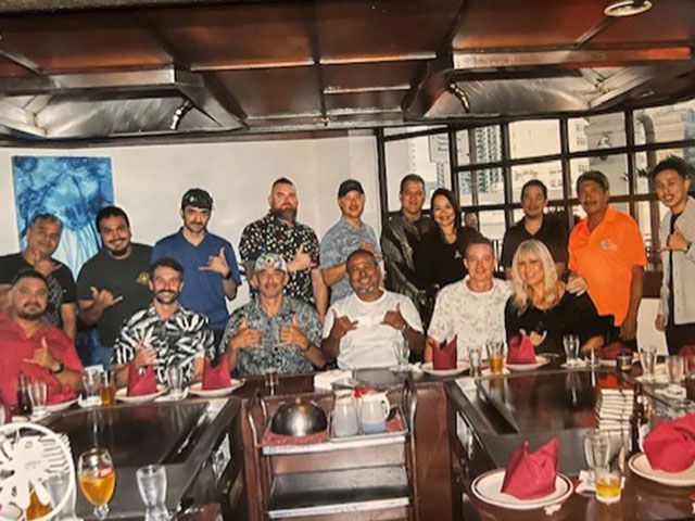 Group of people gathered around a dining table at a restaurant. Some are smiling, holding up shaka signs.