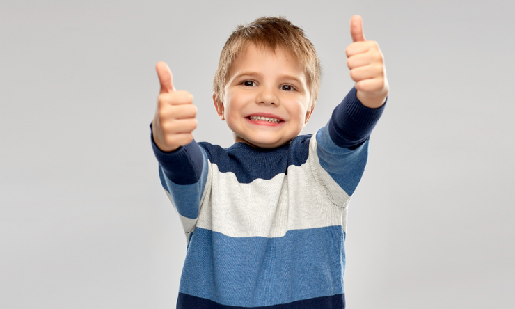 Smiling boy with blond hair giving double thumbs up, wearing a blue and white striped sweater.