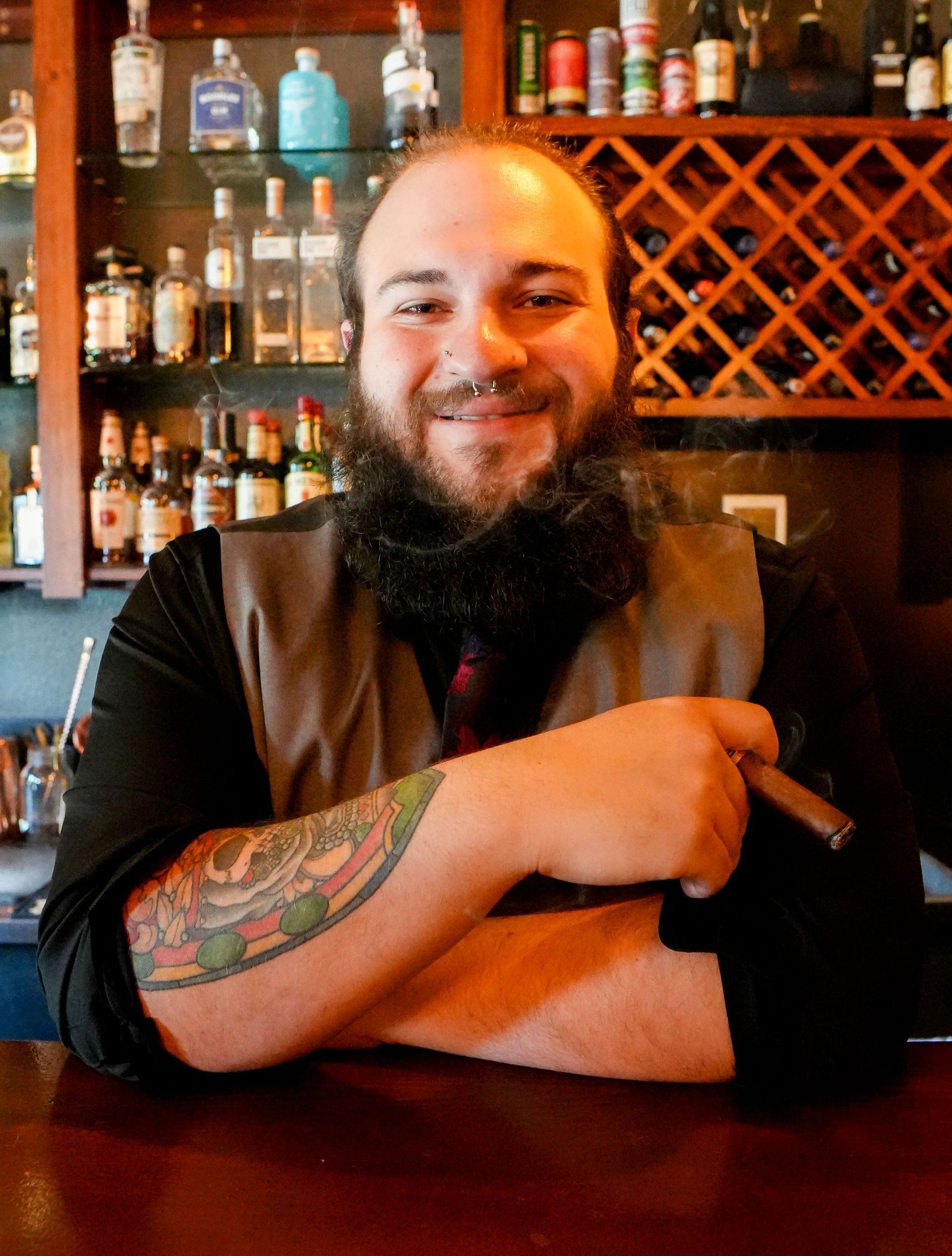 A man with a beard is standing in front of a shelf full of alcohol bottles