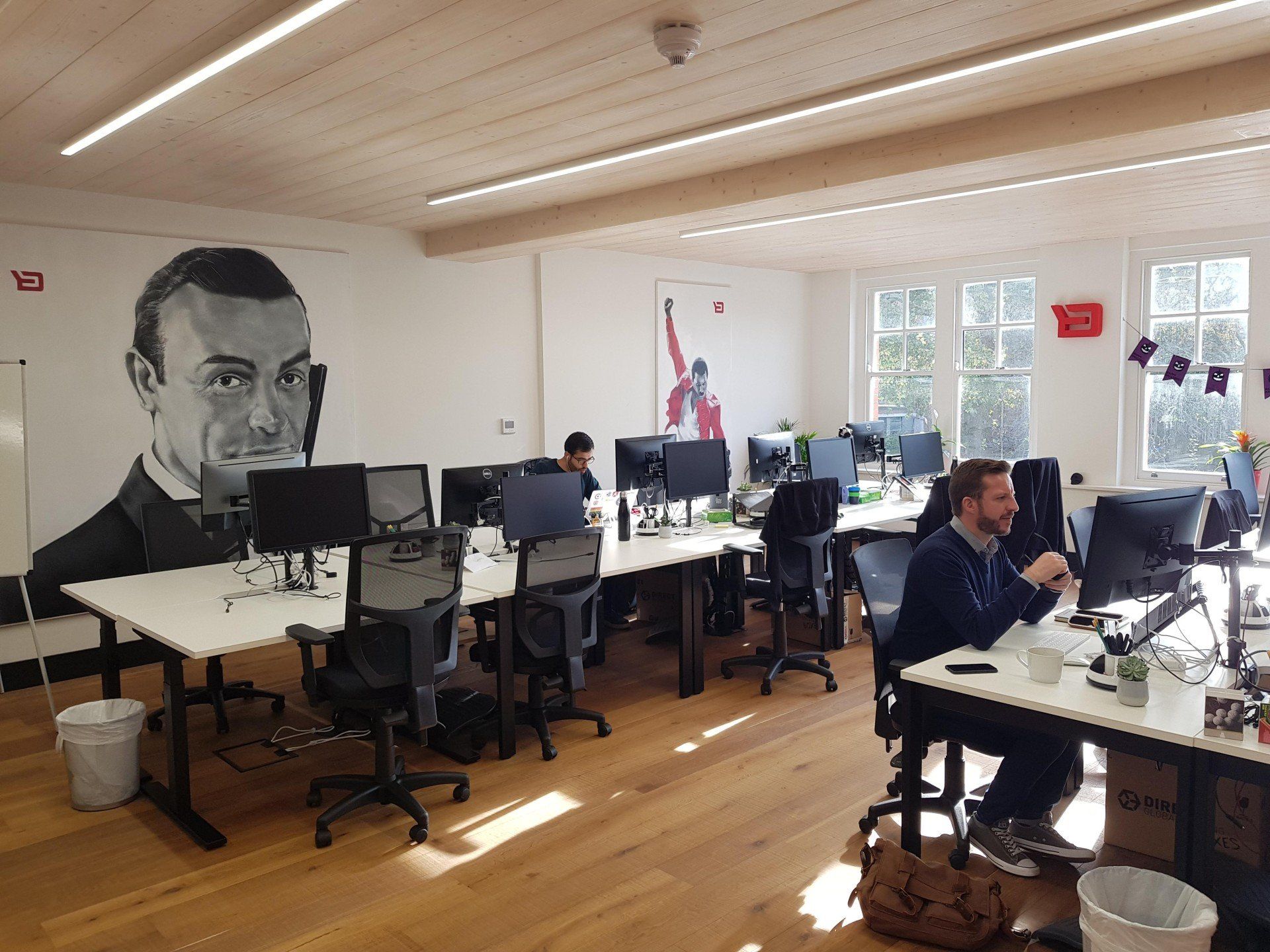 A man is sitting at a desk in an office with a painting of james bond on the wall.