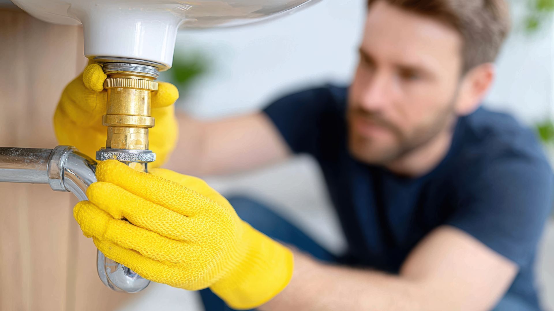 Man wearing yellow gloves repairing plumbing under a sink, focusing on tightening a pipe connection