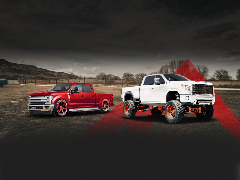 Two customized trucks, one red and one white, parked outdoors. The white truck is lifted with large orange wheels. | Wind River Auto Diesel