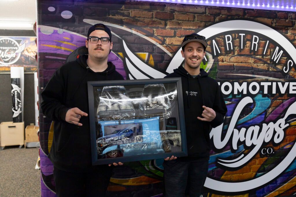 Two men are holding a framed picture in front of a graffiti wall. | Wind River Auto Diesel