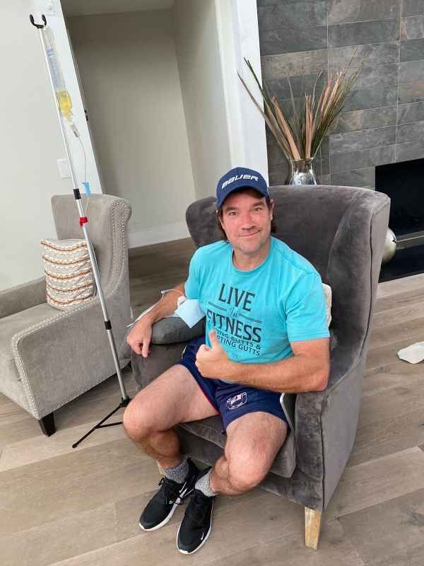 A person wearing a teal t-shirt and baseball cap sits in a chair giving a thumbs-up while receiving an IV infusion.