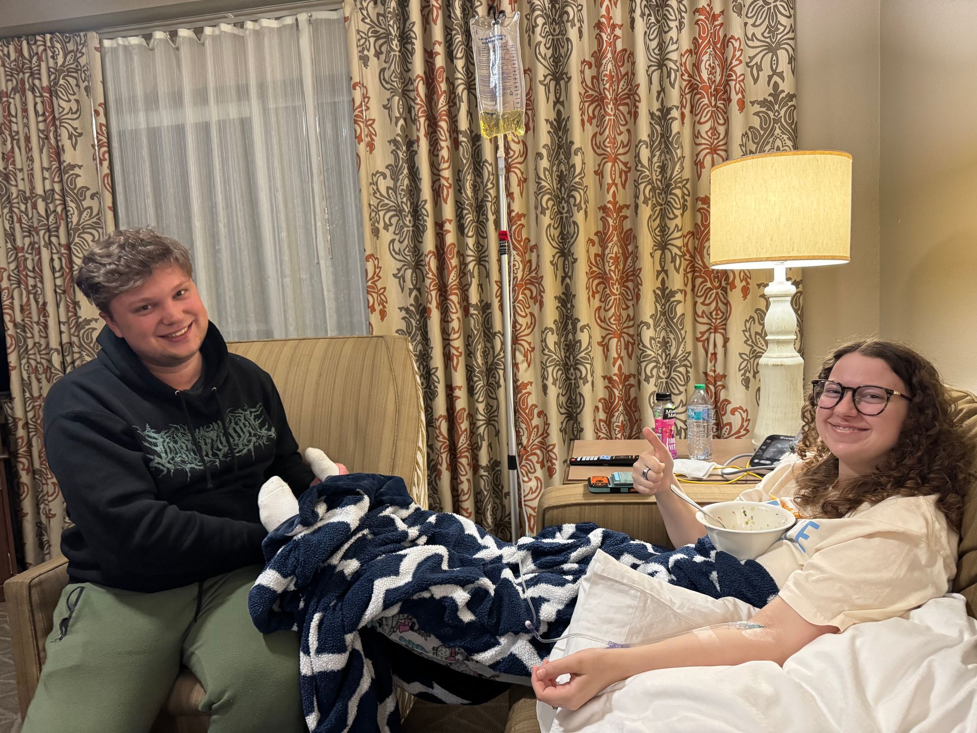 Two people in a room with a hospital IV stand; one sits in a chair, the other gives a thumbs up from a bed under blankets.