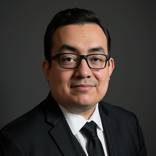 Man wearing glasses and a suit, smiling, against a gray backdrop.