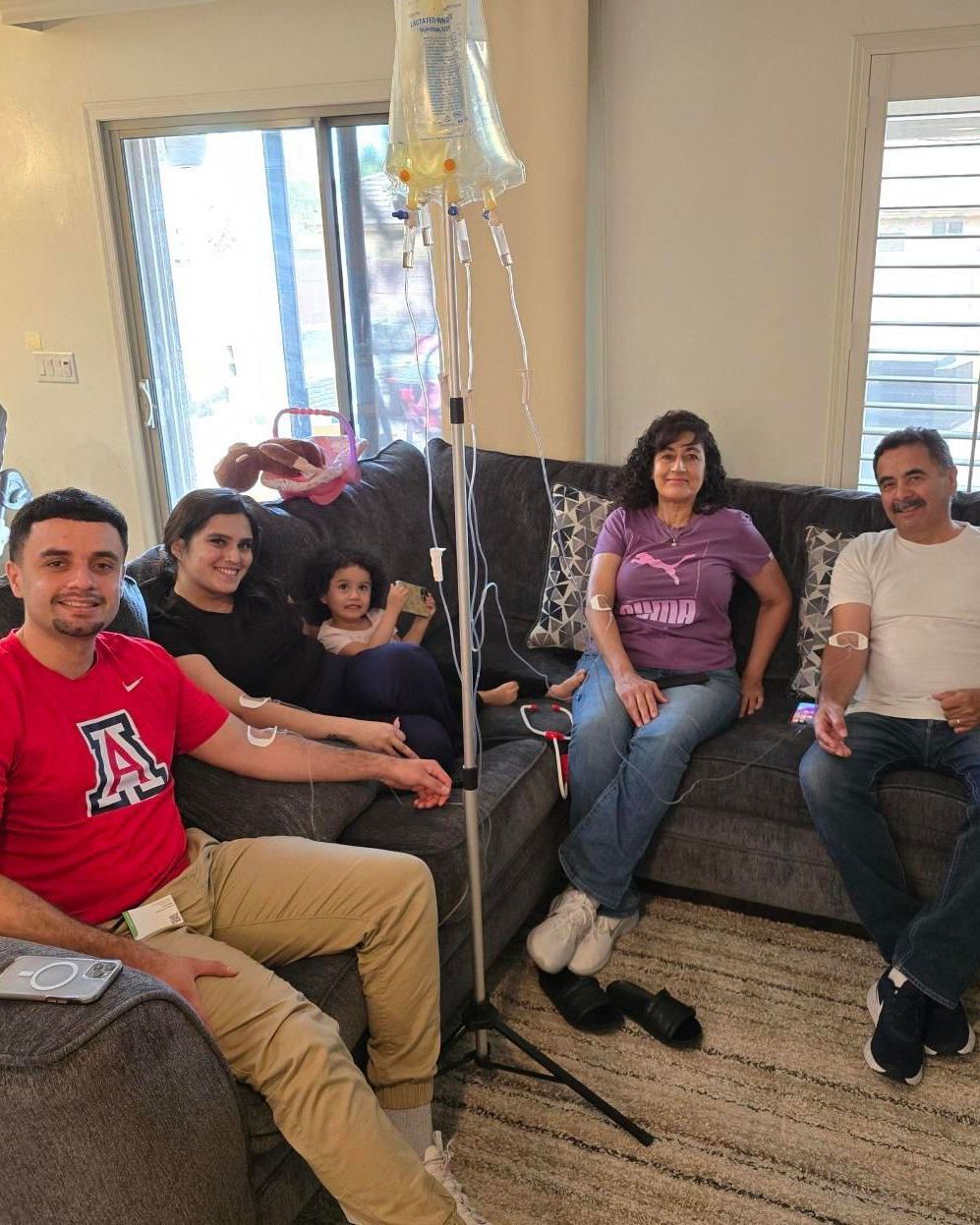 A family sits on a gray sofa in a living room, with three people connected to an intravenous drip on a metal stand.