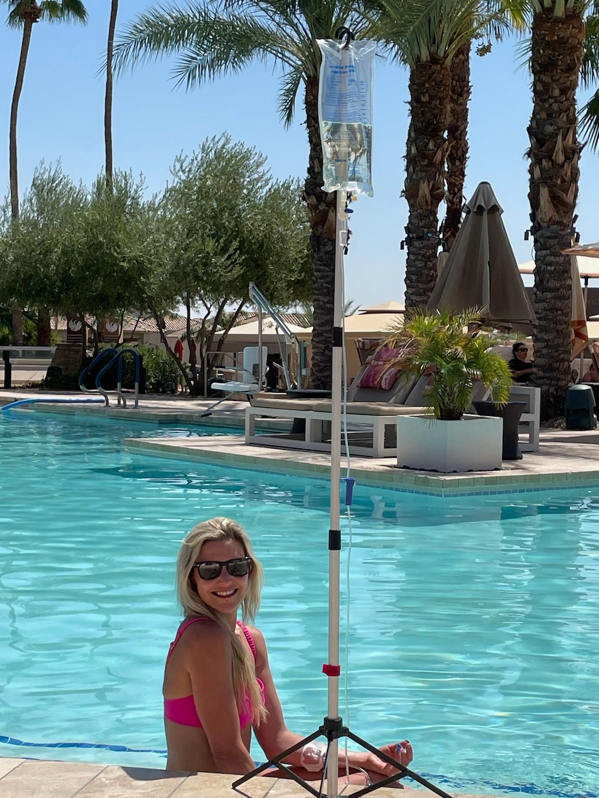A person in a pink swimsuit smiles by a swimming pool next to an IV drip bag on a portable stand.