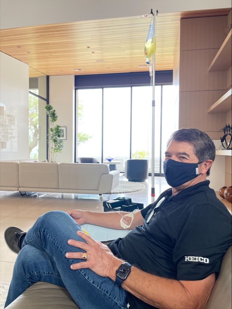 A person in a black mask and shirt sits on a sofa, receiving an IV drip in a bright, modern living room.