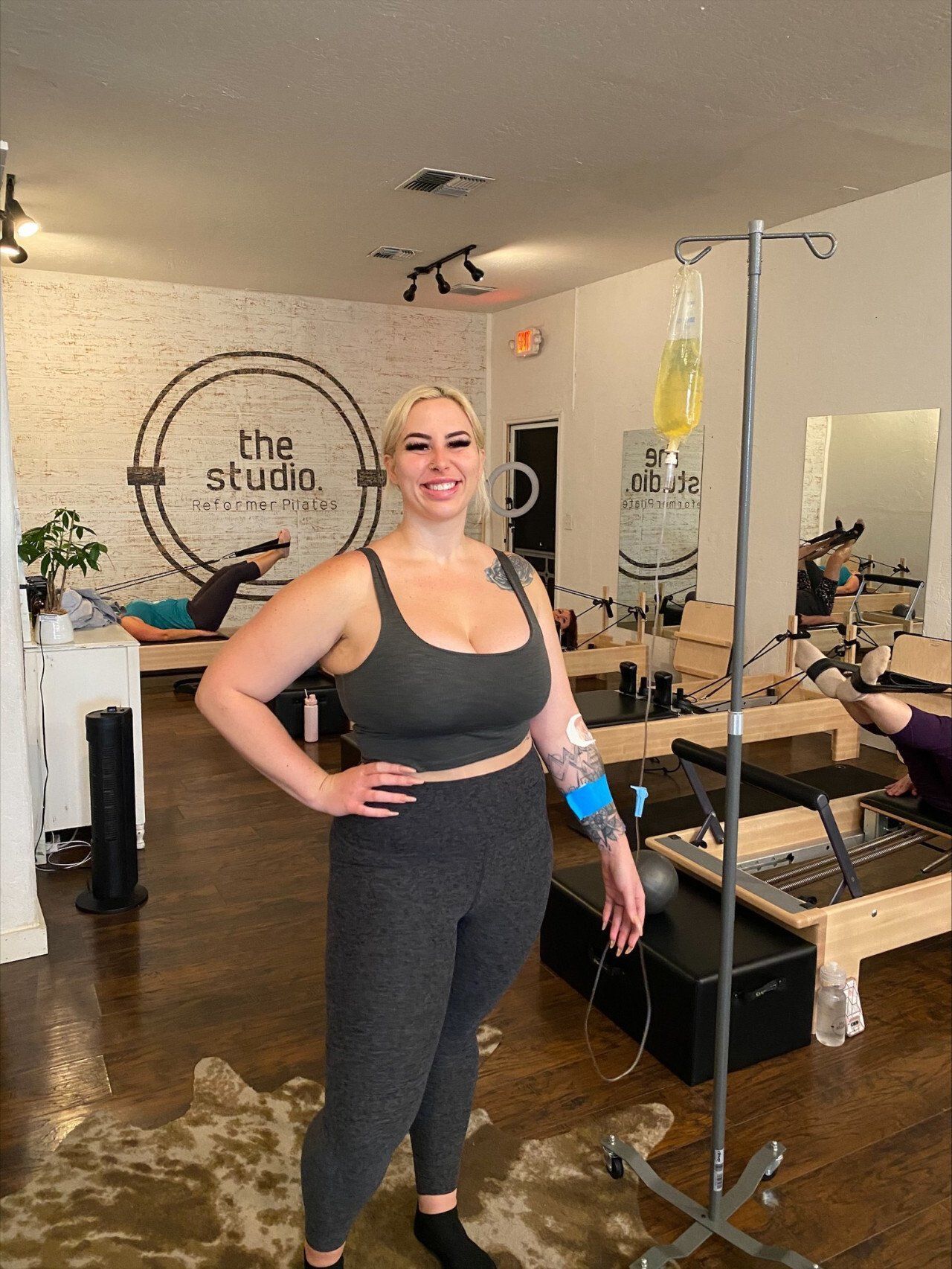 A person smiles while standing next to an IV drip stand inside a fitness studio with Pilates equipment.