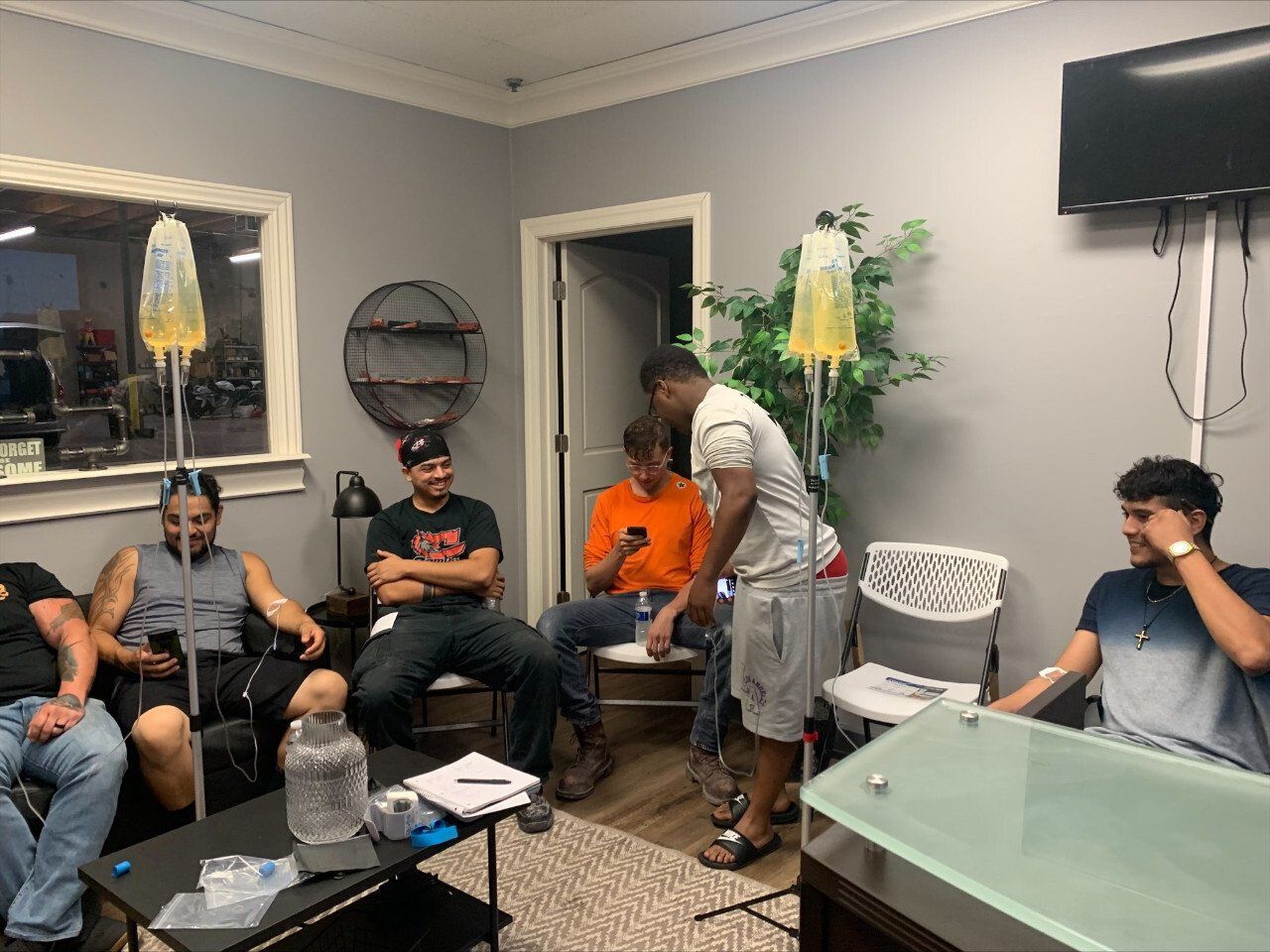 A group of people sitting in a clinic room, some receiving intravenous fluids from IV bags on portable stands.
