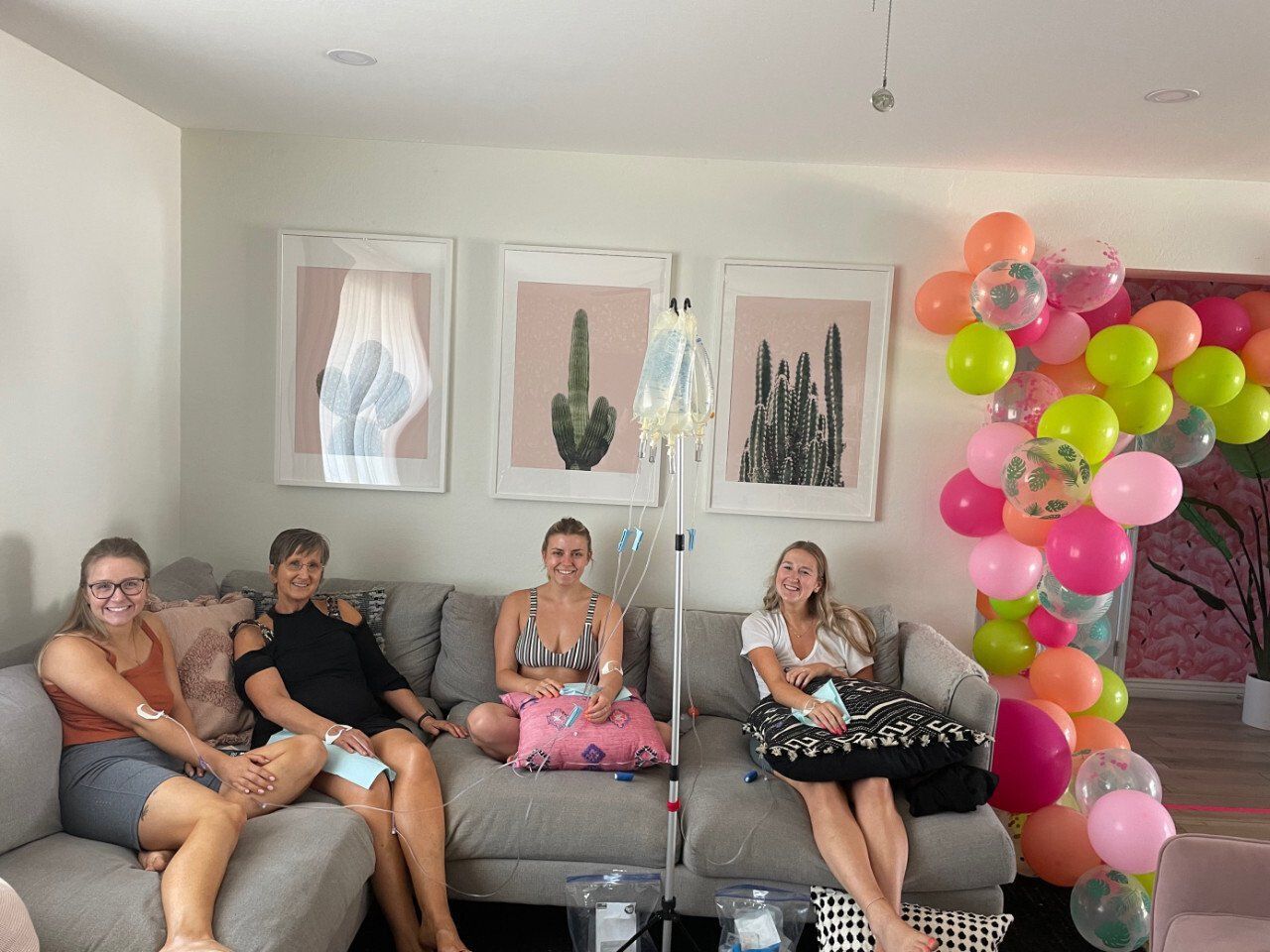 Four people sit on a couch in a room decorated with balloons and framed cactus art, connected to IV bags on a stand.