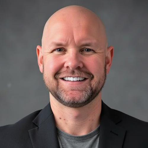 Bald man with a trimmed beard smiles at the camera, wearing a blazer over a gray shirt against a gray background.