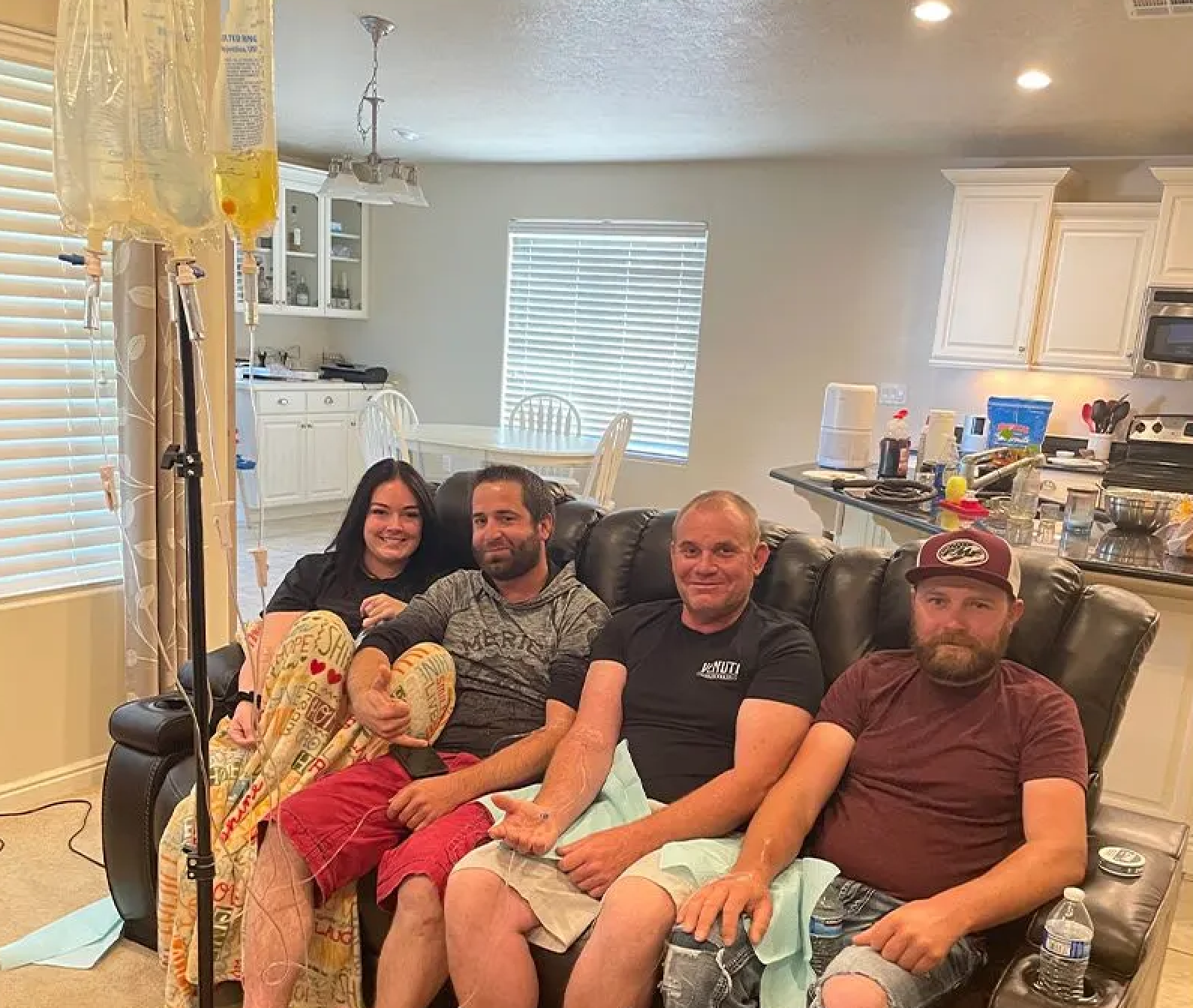 Four people sit on a leather sofa in a kitchen, one with an IV pole nearby.