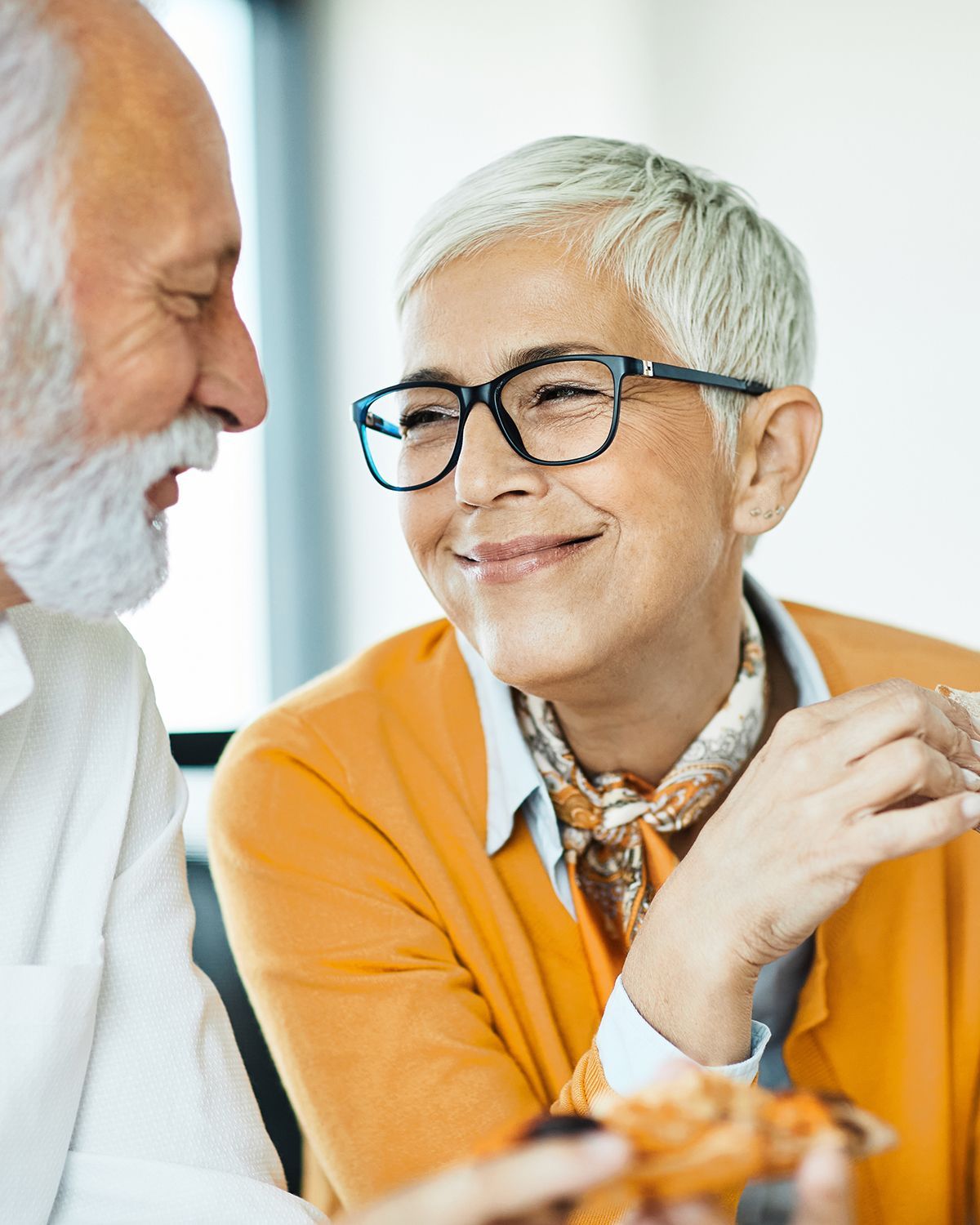 Two individuals with white hair smiling at each other while sharing a slice of pizza.