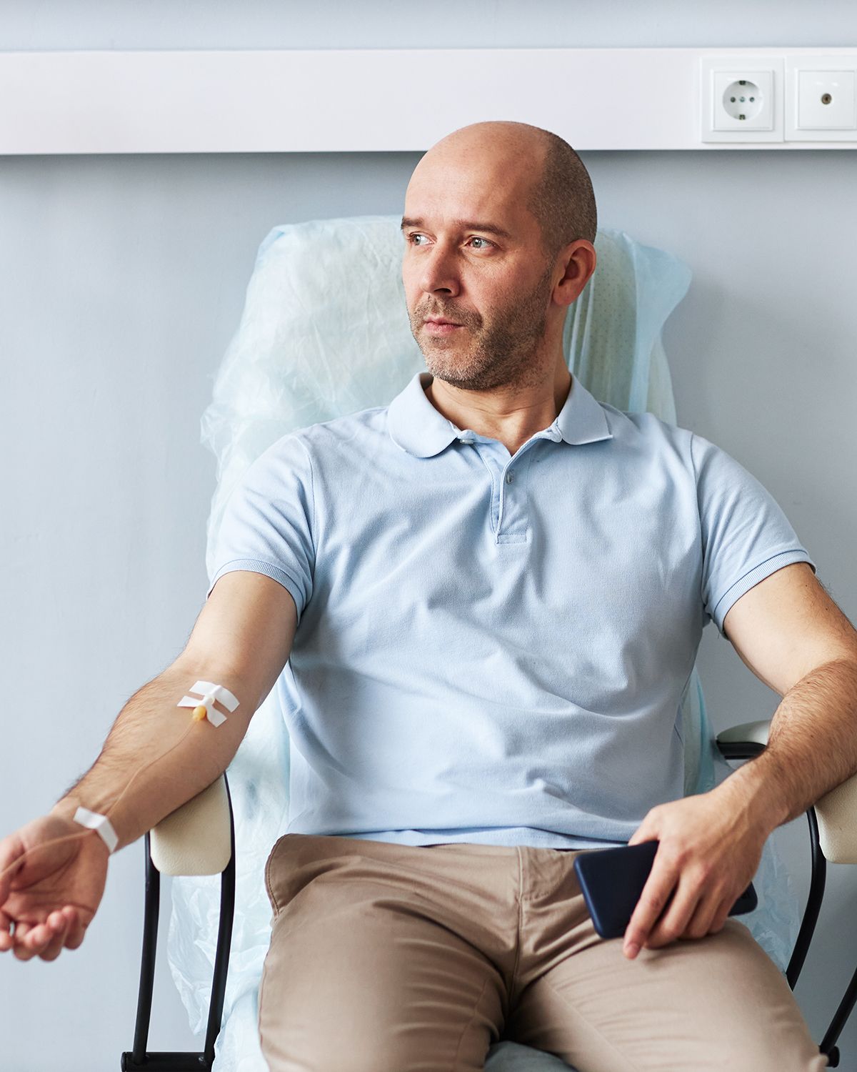 A person sits in a clinic chair with a bandage on their arm after a blood draw, holding a smartphone.