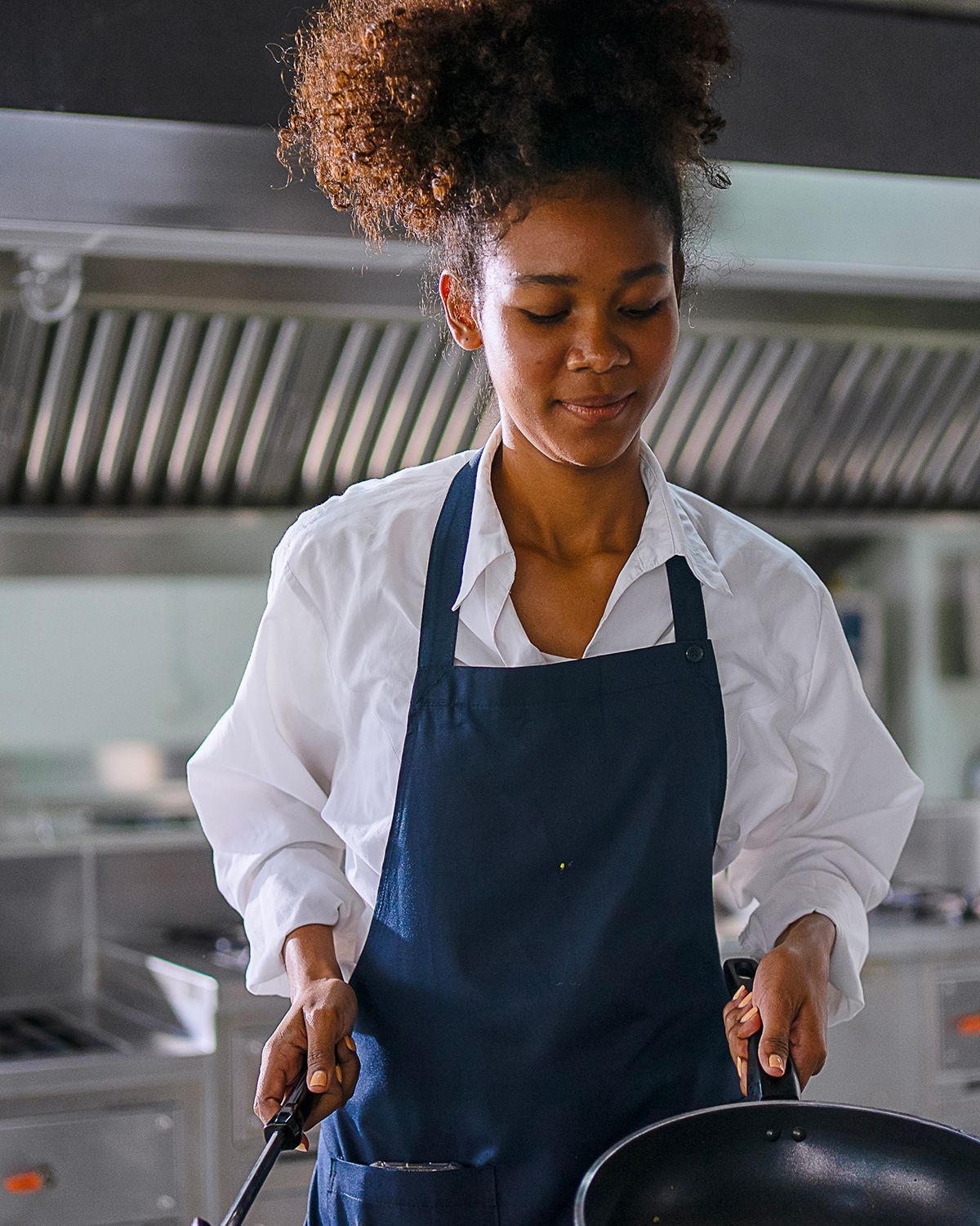 A person wearing a white shirt and dark blue apron prepares food in a professional stainless steel kitchen.
