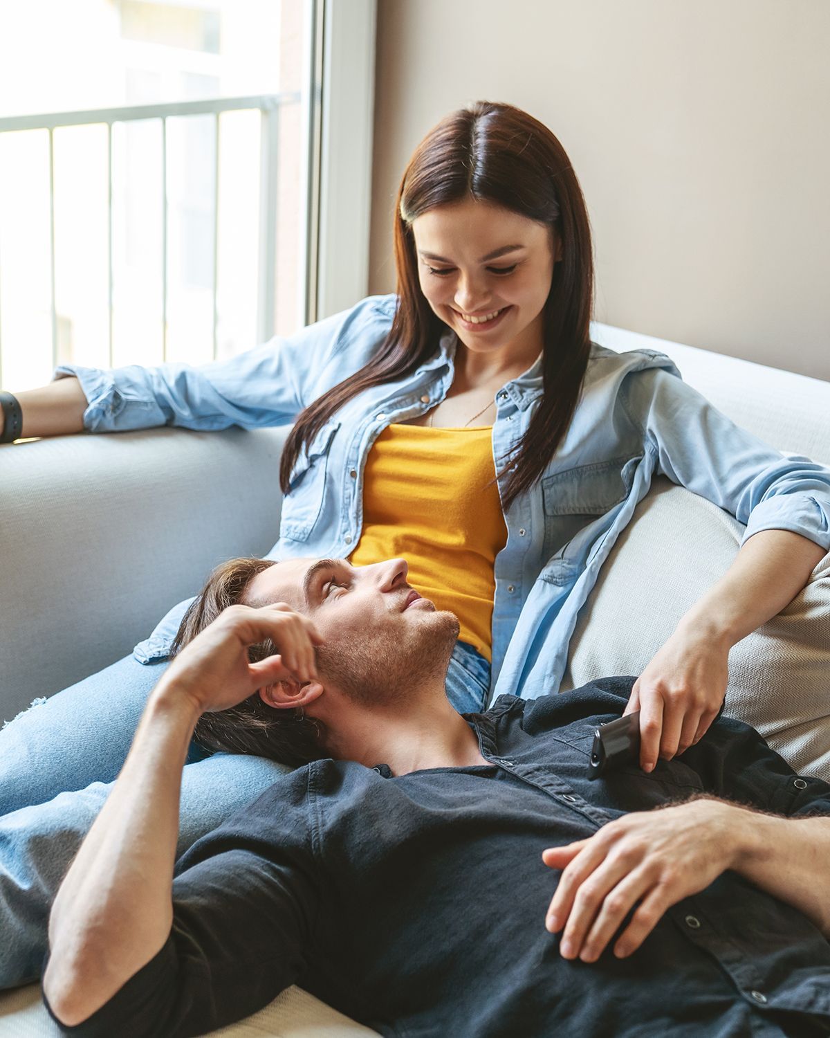A smiling person sits on a sofa while another person lies with their head on their lap, looking up toward them.