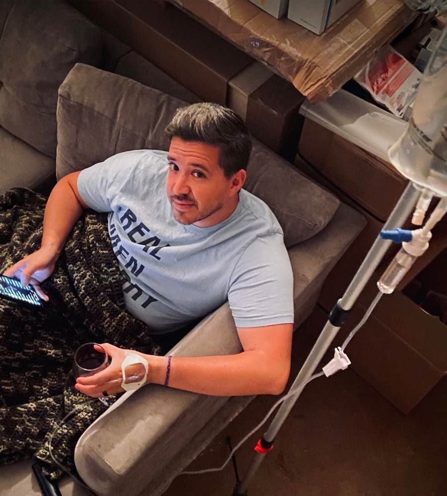 Man on couch with IV drip, holding phone and drink, in a room with boxes.