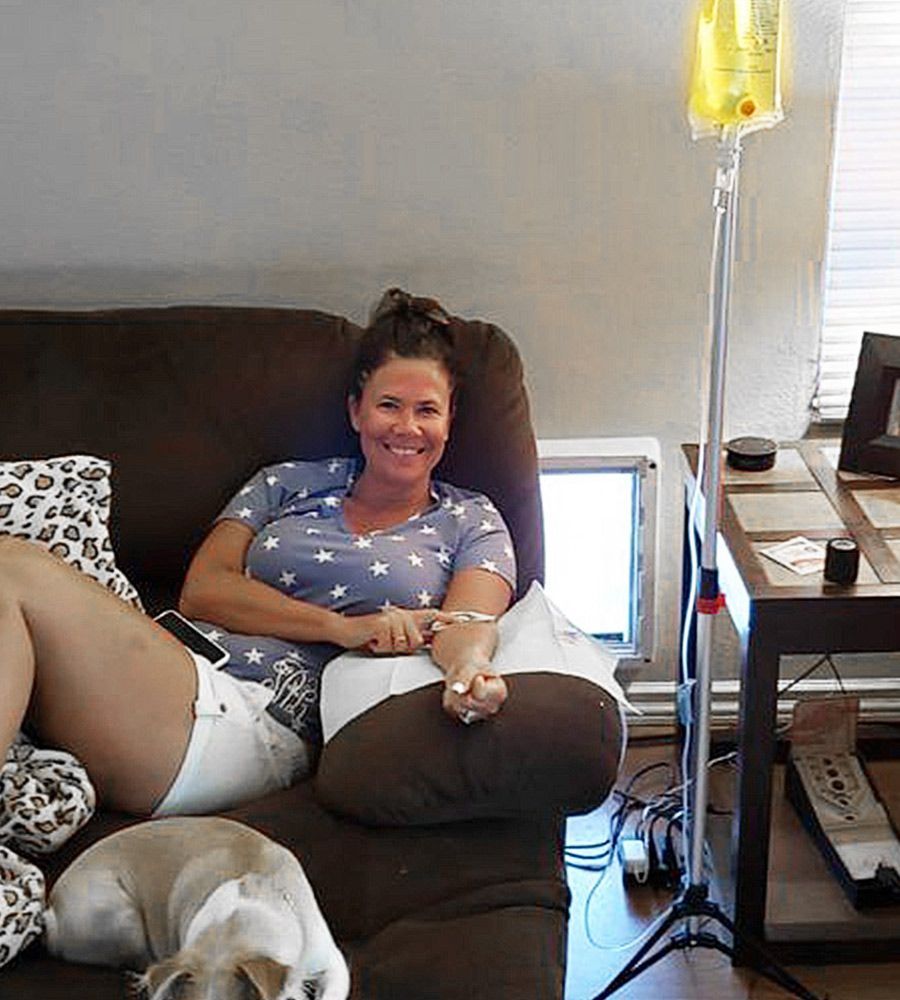 Woman receiving IV fluids on couch, smiling. Dog nearby. IV pole and side table in the room.