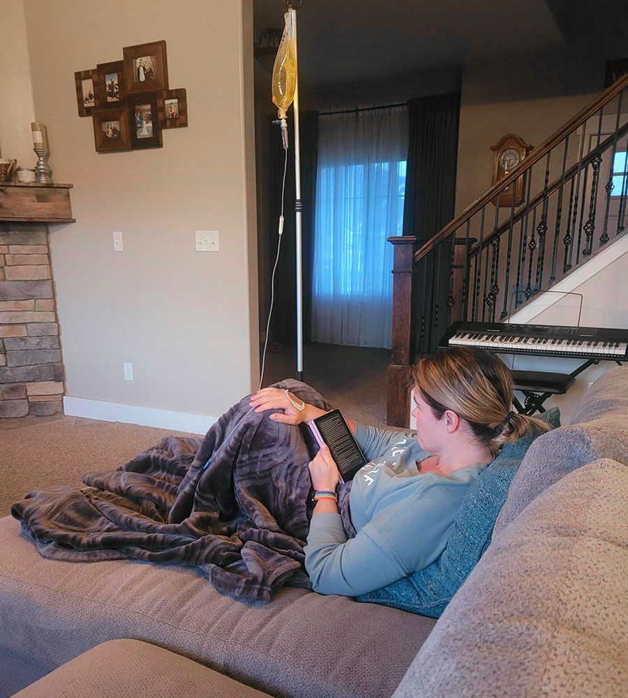A woman receiving at-home IV therapy while resting on a couch and reading a digital book.